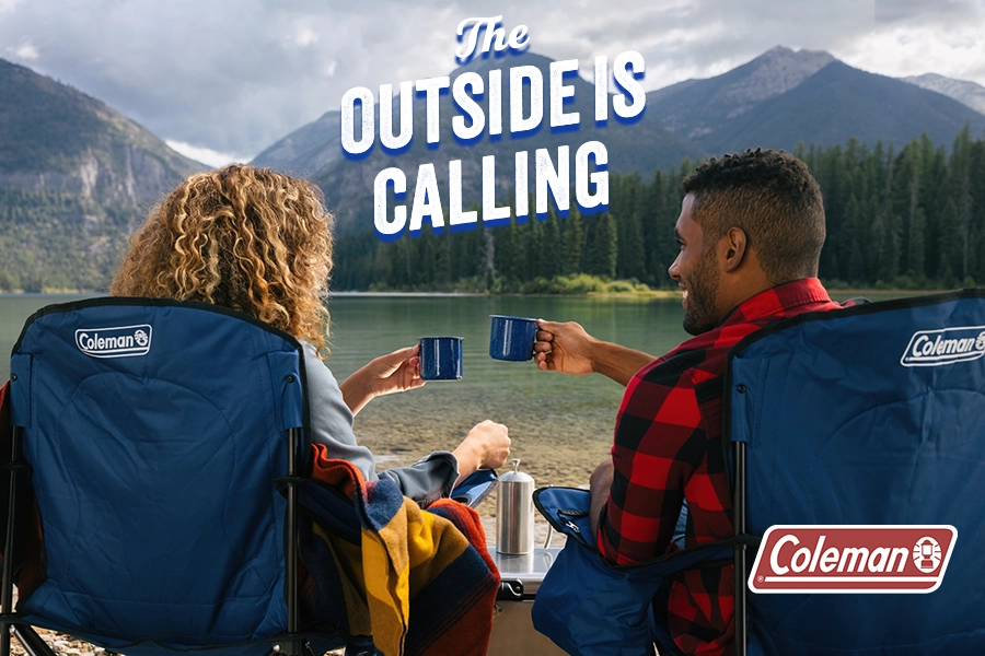 The OUTSIDE IS CALLING couple raising their camping mugs of a warm beverage sitting in camp chairs in the outdoors overlooking a mountain lake.